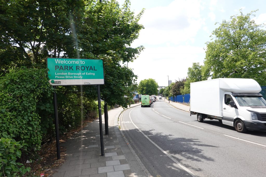Photo shows a sign saying 'Welcome to Park Royal, London Borough of Ealing, Please Drive Slowly'. The sign clutters a narrow footway. There is a narrow paint-only cycle lane on one side of a busy but narrow road.