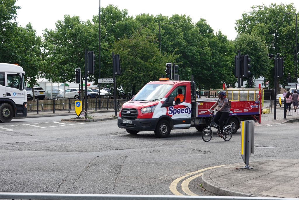 Person riding a Brompton negotiating a busy junction with HGVs and van traffic.