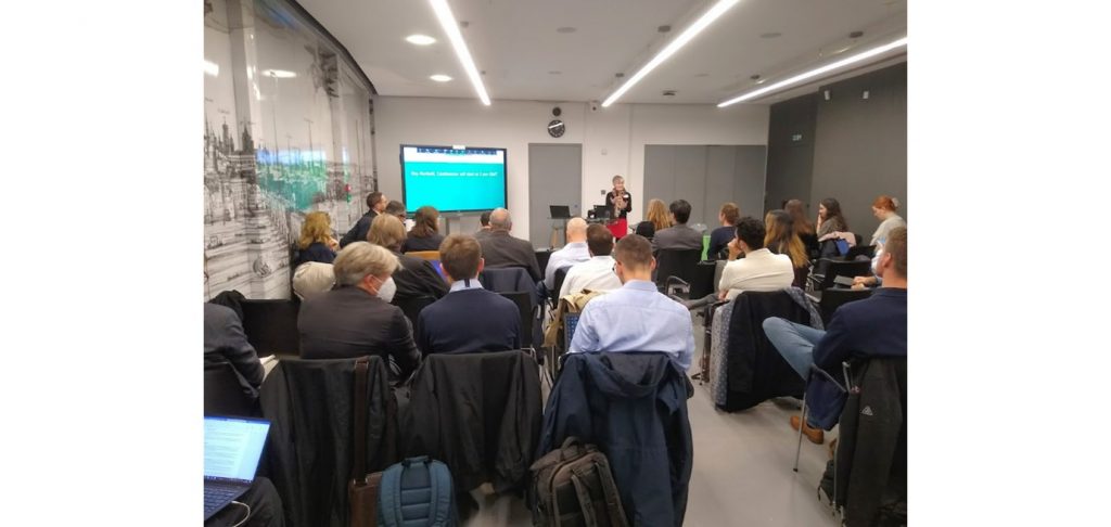 A photo of conference delegates at the ReVeAL Cities conference, London sat watching Bonnie Fenton of Rupprecht Consult talking in front of a presentation on a TV screen.