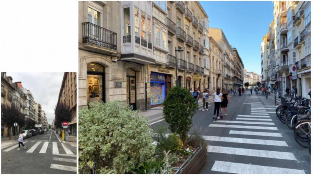 A photo of a street within the San Antonio superblock in Vitoria-Gasteiz. The photo shows planters and plants in the middle of the street, white lines painted on the street and people walking on the newly pedestrianised street.