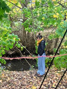 A person wearing headphones standing next to a river and amongst some trees. She is wearing a yellow scarf, a black jacket and blue jeans.