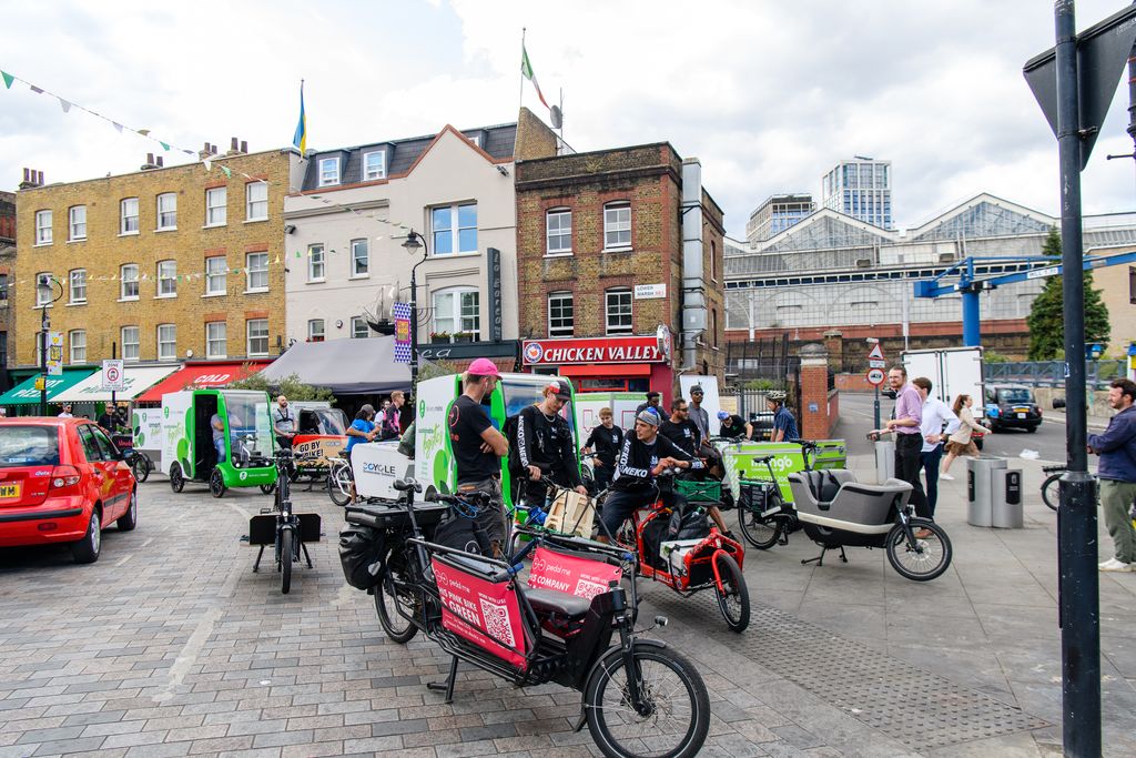 A photo of a street scene. In the background are shops. In the foreground are a collection of different styles of cargo bikes and their riders.