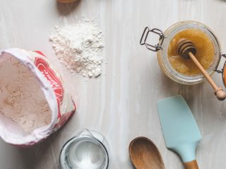 A worktop with various baking implements on top - a wooden spoon, spatula, eggs, flour, jug of water and a jar of honey and honey turner.