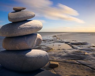 Stack of 5 pebbles to the left of the photo, which is looking out on a coast line horizon during the day. There is blue sky and sunshine