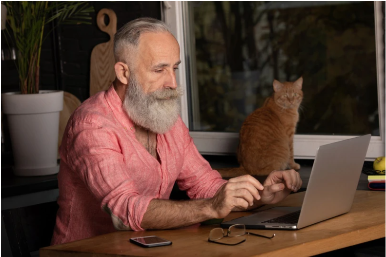 Man with a pink shirt on sitting at a desk in front of an open laptop with an orange cat in the background.