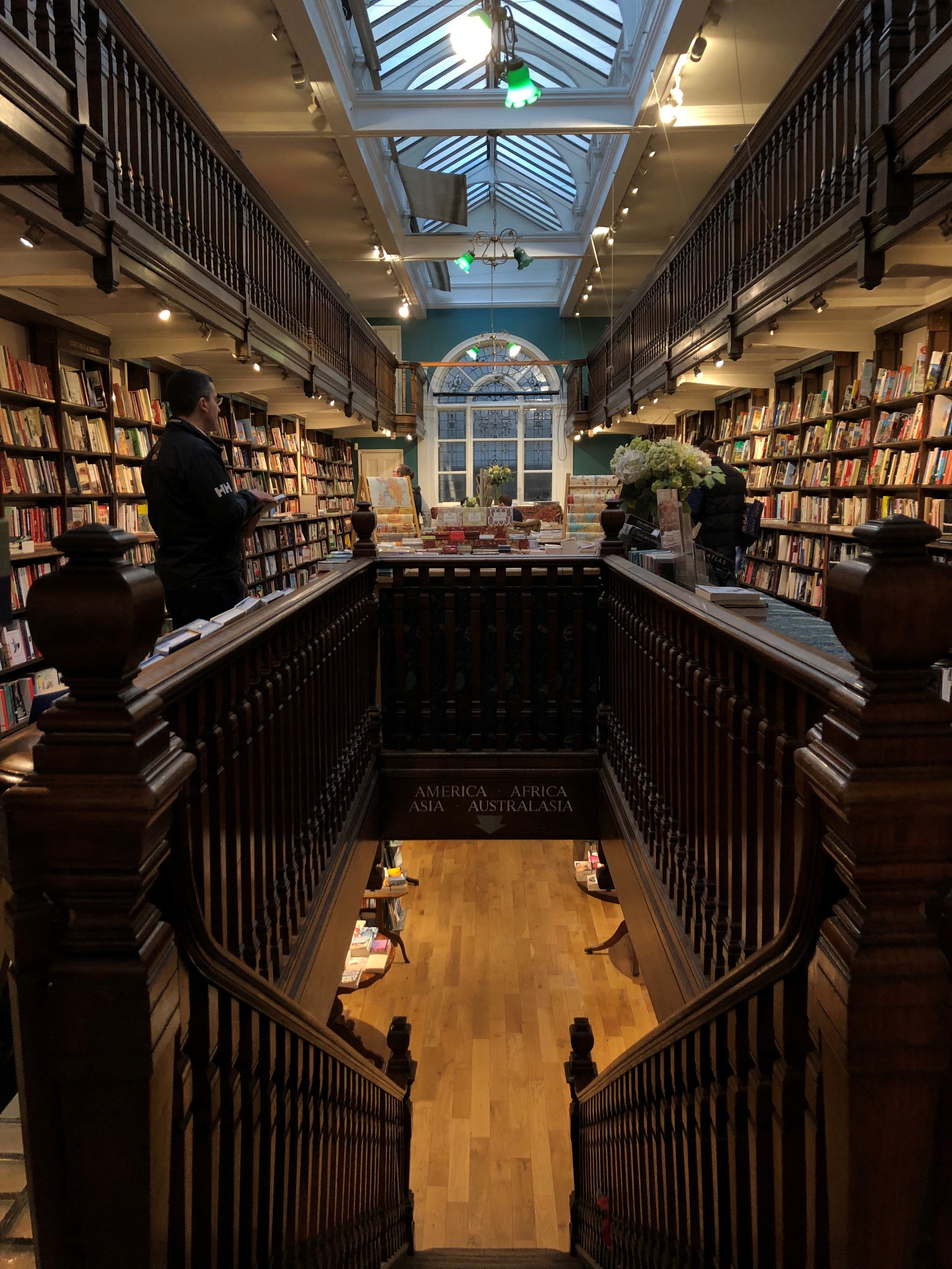 Daunt Books Marylebone bookshop gallery book-lined shelves, sky[light, wooden carved banisters, stairs leading down to lower gallery, green lamps