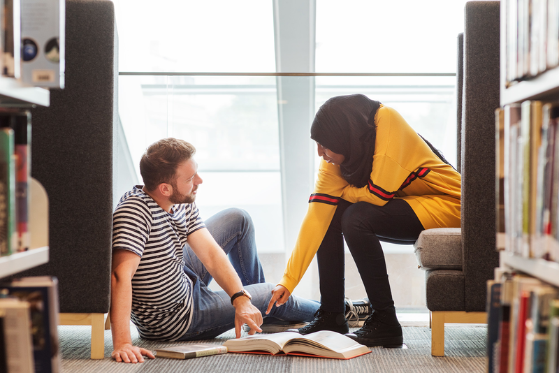 Two students sitting on the floor of the library looking at books together