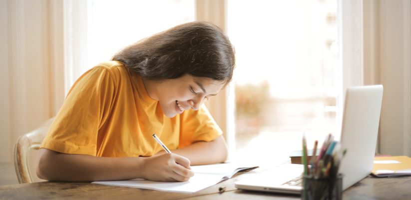 Student in yellow t-shirt writing in a notebook in front of a laptop.