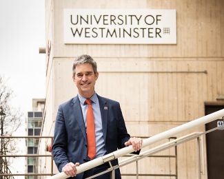Professor Peter Bonfield wearing a navy suit and orange tie. He is standing on stairs holding the railing in front of a sign that reads UNIVERSITY OF WESTMINSTER