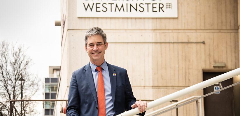 Professor Peter Bonfield wearing a navy suit and orange tie. He is standing on stairs holding the railing in front of a sign that reads UNIVERSITY OF WESTMINSTER
