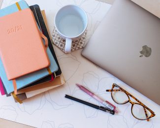An aerial image of a mug, laptop, notebooks, pens and glasses on a desk