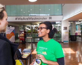 Student ambassador wearing green 'Here to Help' t-shirt advising a guest at an open day