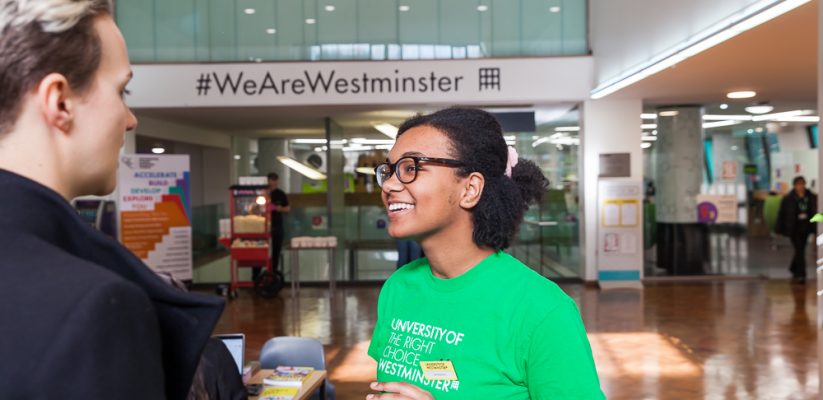 Student ambassador wearing green 'Here to Help' t-shirt advising a guest at an open day