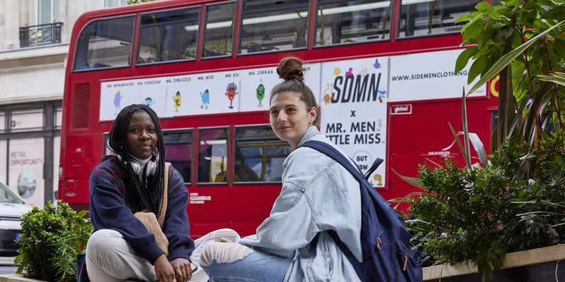 two-female-students-sitting-outside-regent-campus-with reb-bus-in-background