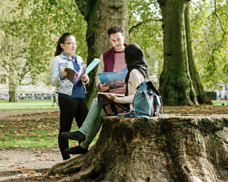 A male and two female students in a park or square, one of them sitting on a tree stump, talking, smiling, holding various prospectuses; trees visible in the background, close-up