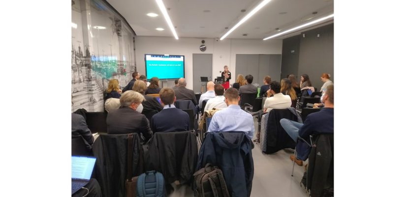 a photo of conference delegates at the ReVeAL Cities conference, London sat watching Bonnie Fenton of Rupprecht Consult talking in front of a presentation on a TV screen.